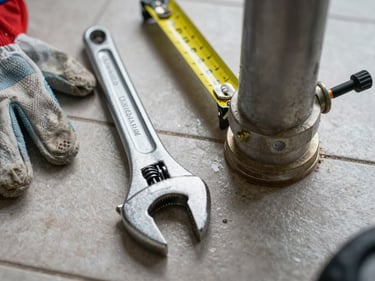 Overhead close-up of a plumber's wrench and tape measure laid on a tile floor beside a pipe joint, cool shop lighting, work gloves nearby, no people visible