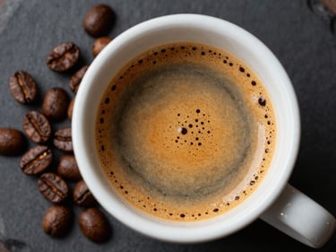 Overhead close-up of a freshly pulled double espresso in a ceramic cup, golden crema surface, dark roast beans scattered beside it on black slate, studio strobe from the left