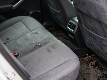 Close-up of a car interior with waterline stains on the seat fabric and mud residue on the floor mat, natural daylight through the window, functional documentary framing