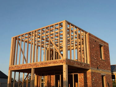 Structural timber frame of a single-storey residential extension mid-build, open to the sky, brickwork rising on one side, golden hour light raking across the timbers