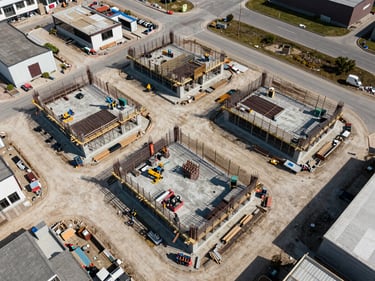 Overhead orthographic view of a commercial construction site mid-build, concrete foundations and steel framing visible from directly above, site access roads and staging areas in natural daylight