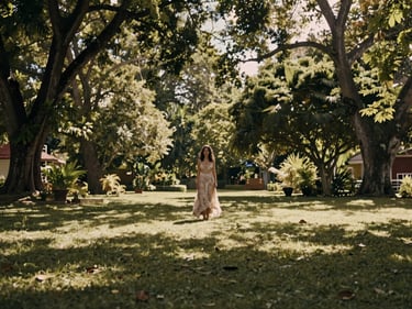 Wide environmental frame, young woman in a flowing dress standing in a shaded garden in Montería, soft north-facing daylight filtering through tree canopy, subject off-center to the left, generous negative space on the right, warm muted tones, no harsh shadows