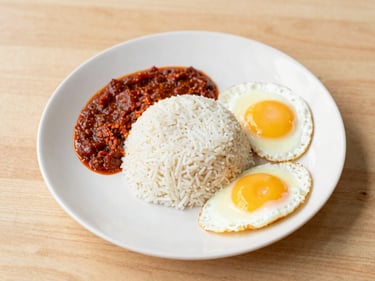 Overhead shot of a full Malaysian breakfast plate with nasi lemak, fried egg, and sambal, on a wooden table, bright natural light, soft yellow accents.