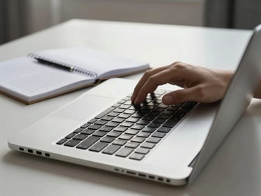 Hands typing on a laptop on a clean desk, a spiral notebook open beside it with written notes, natural window light, sharp detail on the keyboard keys, minimal background
