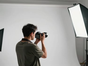 Wide environmental shot of a photographer from behind, holding a camera and adjusting settings while standing in a lit studio bay, softbox light visible on the right, white seamless behind the subject area