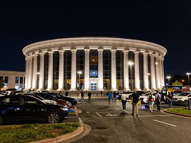 Wide environmental shot of The Forum's iconic circular exterior at night, event lighting spilling onto the parking perimeter, crowd moving toward curb pickup zones, real ambient streetlight, dynamic angle from ground level