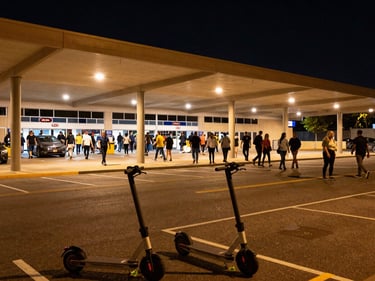 Wide environmental shot of Kia Forum parking perimeter at night, crowd streaming from the arena exit toward the street, overhead parking lot lights casting real amber glow, electric scooters staged at the curb in the foreground, dynamic angle from street level