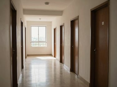 Wide interior corridor of a mid-rise apartment building in Chennai, natural north light flooding through end window, polished floor tiles, numbered apartment doors in a row