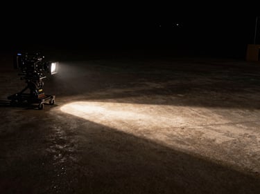 Empty film set at night, practical tungsten fresnel casting a hard diagonal shaft across a dark concrete floor, camera on a dolly in the left third, deep background falling off to black, cinematic medium wide shot