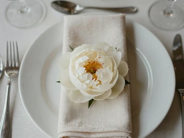 Close-up overhead of a finished wedding table arrangement — folded ivory napkins, crystal glassware, a single white peony at centre, soft diffused overcast light, linen texture visible