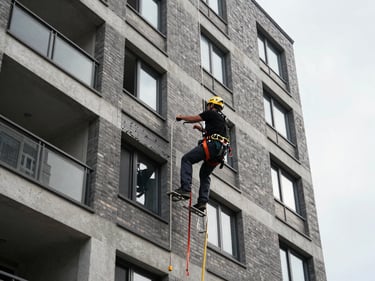 Exterior view of a multi-story residential condominium facade, technician in full safety harness working on upper floor cladding, overcast natural daylight