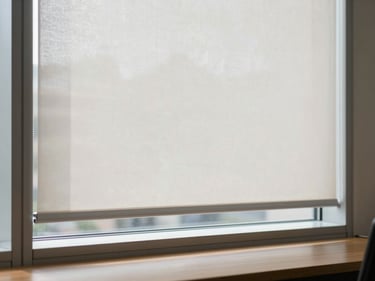 Interior shot of a corporate office window with a fabric roller blind partially lowered, soft natural light filtering through the blind material creating an even wash of light across a wood desk surface below, clean white window frame, no people, overcast daylight