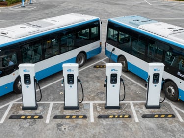 Overhead view of a municipal vehicle depot parking area, four EV charging stations installed in a row, charging cables connected to electric buses, flat grey tarmac, overcast daylight