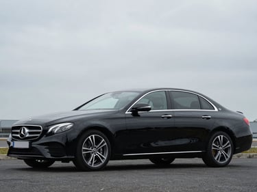 Front three-quarter driver-side profile of a black Mercedes-Benz E-Class, overcast daylight, full vehicle visible, squared frame, no tilt