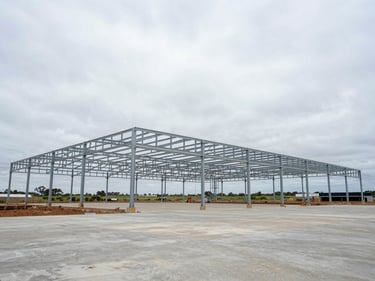 Wide-angle ground-level view of a commercial agricultural facility under construction on the KwaZulu-Natal south coast, steel portal frame rising against an overcast sky, concrete slab visible, no workers posed