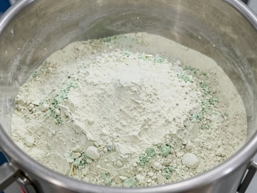 Overhead view of a pharmaceutical powder blending vessel interior, fine white-and-green powder blend uniformly distributed, stainless steel walls, bright facility lighting, no people