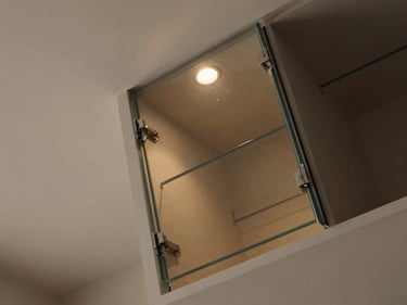Overhead 45-degree view of a built-in glass-fronted storage unit recessed into a residential wall, sharp focus on the frameless glass door hinge and interior shelf edge, warm overhead light, muted neutral wall finish