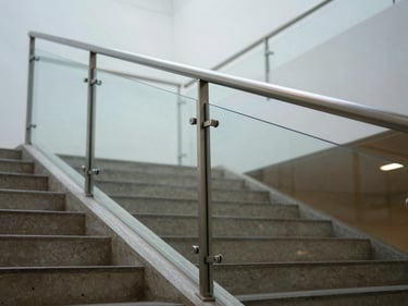 Wide-angle view of a frameless structural glass balustrade at the edge of an interior staircase, sharp focus on the stainless point-fix fitting at mid-span, cool diffuse daylight from above, warm concrete stair treads visible below
