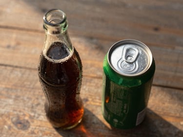 Overhead close-up of a glass soda bottle and a can on a worn wooden surface, golden afternoon light from the left, condensation visible on the bottle