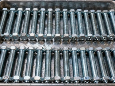 Wide overhead shot of sorted galvanized fasteners — hex bolts, carriage bolts, lag screws, and machine screws arranged in rows on a steel tray, warehouse overhead lighting, extreme texture detail on threads and zinc coating, no human presence