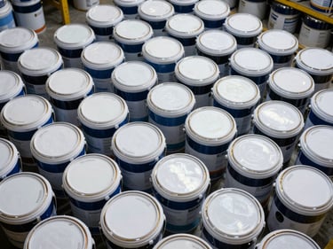 Overhead cataloging shot of stacked paint buckets and primer drums in a warehouse, product labels and sheen codes visible on lids, organized rows showing inventory depth, diffused industrial lighting, no human presence