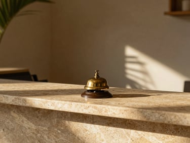Minimalist boutique hotel reception desk with a warm stone counter, a brass bell, soft shadows, tropical sunlight, warm editorial style