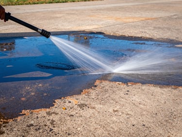 High-pressure water stream cleaning an oil-stained driveway, sharp contrast between clean and dirty, direct Texas sunlight, saturated blue and orange tones.