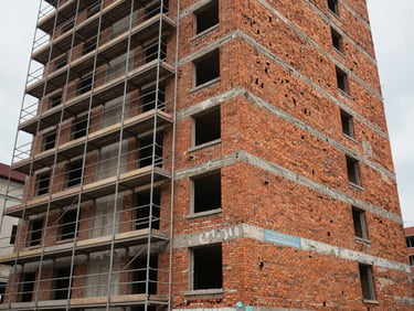 Exterior facade of a mid-rise residential building under construction, scaffolding on left third, finished brick cladding visible on right, overcast natural daylight, street-level perspective