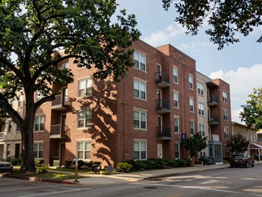Wide street-level exterior shot of a six-story brick multi-family apartment building in Austin, Texas, natural afternoon daylight, mature street trees in foreground, neighborhood context visible