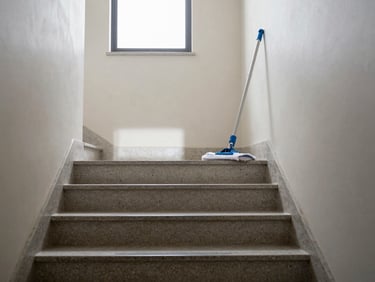 Eye-level view down a clean residential stairwell, polished stone steps, mop leaning against the wall, natural light from a landing window, no people