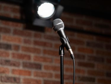 Close-up of a microphone stand on a dimly lit comedy club stage, harsh overhead spotlight, raw brick wall behind, no performer visible