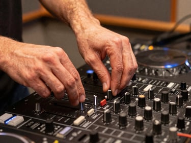 Close-up of a DJ's hands on a mixing board inside a small radio studio, warm tungsten light, analog dials and sliders in sharp focus, candid mid-broadcast moment