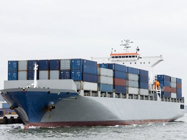 A high-angle shot of a modern cargo ship loaded with blue and gray shipping containers at an international port, overcast daylight