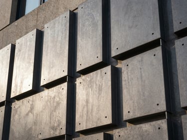 Close-up of a precision-cut steel architectural facade panel installed on a building exterior, afternoon side-light raking across the perforated surface pattern, sharp shadow lines defining the cut geometry, warm grey steel tones against concrete