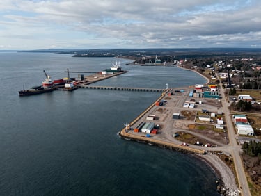 High-altitude aerial view of a raw deep-water access plot on the Cape Breton coastline, adjacent to active industrial piers, overcast daylight.
