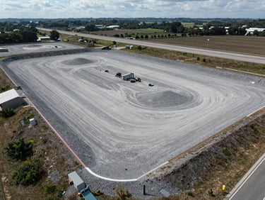 Drone view of a cleared gravel laydown yard with marked boundaries, adjacent to a major highway corridor, high-clarity overcast daylight.