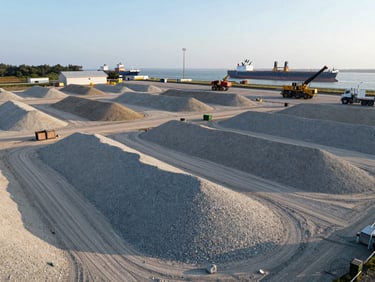 Drone perspective of a massive gravel-packed laydown yard near maritime shipping lanes, heavy equipment visible in background, crisp morning light.
