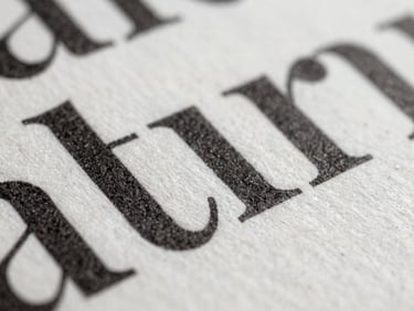 A macro close-up of a heavy cotton paper page, deeply debossed black ink typography, soft north-facing daylight casting subtle shadows across the texture.