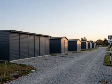 Wide environmental shot of an open yard with anthracite-painted storage units and a gravel access road, a regional cycling route signpost visible at the far right edge, soft late-afternoon light, no people
