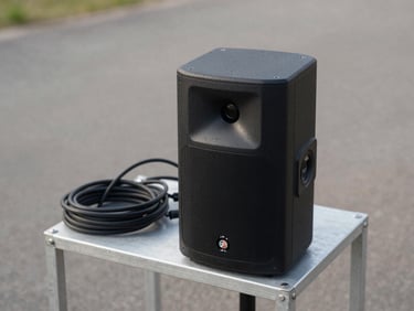 Close-up of a compact PA speaker on a metal stand positioned outdoors on a paved surface, cables neatly coiled beside it, neutral daylight, equipment-focused framing with no people