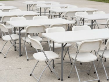 Rows of white folding chairs and rectangular trestle tables arranged in formation on a clean concrete surface, neutral daylight, wide environmental framing showing the full setup