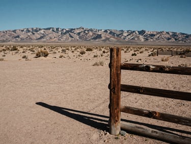 35mm documentary style photograph of a weathered wooden ranch fence post in the foreground under harsh desert sun, vast New Mexico plain stretching to mountains on the horizon, deep shadows, high contrast.