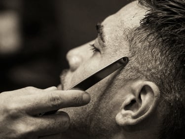 Macro shot of a barber's hand holding a sharp straight razor, precise lines on a client's cheek, dramatic side-lighting, black and white filter, 35mm film.