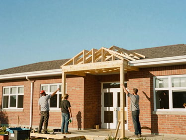 Local builders raising a new timber roof truss on a brick schoolhouse, bright blue sky, warm natural sunlight.
