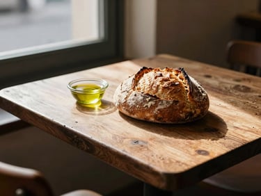 A beautifully styled rustic table at an artisanal restaurant, soft daylight streaming through a window, a fresh sourdough loaf and olive oil.