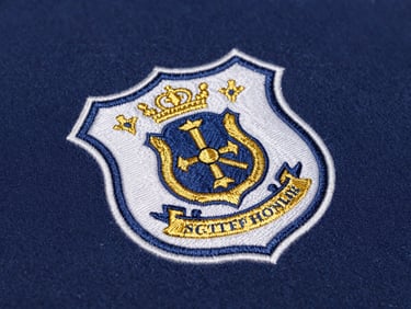 Macro shot of a finely embroidered school crest in gold and blue on a navy fabric swatch, sharp focus on thread detail, clean lighting.
