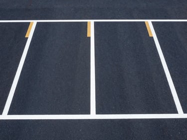 Symmetrical overhead drone shot of freshly applied, razor-sharp white and safety-gold parking lines on dark asphalt. Bright midday sun casts high-contrast, crisp shadows, emphasizing the flawless layout alignment.