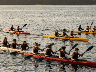 Students kayaking on a serene lake during a school trip, warm golden-hour sunlight, active, candid, wide environmental shot.