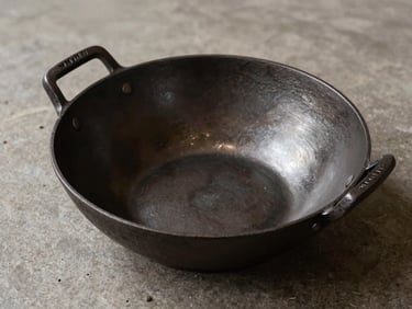 Clean overhead B2B product specification shot of a raw cast iron wok on a neutral stone backdrop, deep charcoal tones, 35mm.