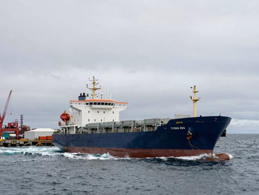 Wide environmental shot of a cargo vessel at anchor in a grey harbour, Southern Ocean swell visible beyond the breakwater, industrial port infrastructure to the left third